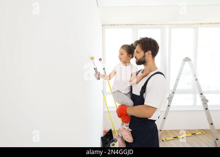 Sorridente ragazza di 5 anni tiene un rullo di vernice nelle sue mani. Il bambino aiuta suo padre, che la tiene, vicino al muro, nella riparazione. Foto Stock