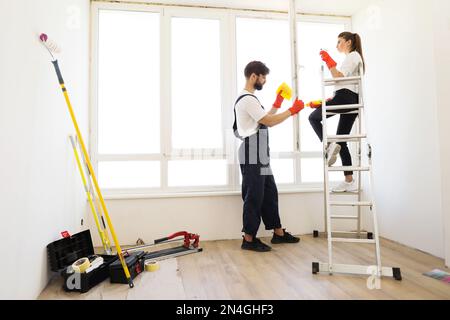 Giovane famiglia di due, bella donna seduta sulla scala e bearded faccende domestiche dell'uomo, lavano la finestra nella loro casa con l'agente di pulizia e gli stracci gialli di pulizia della casa. Foto Stock