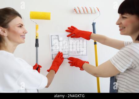 Vista ritagliata di belle giovani donne caucasiche felice fa riparazioni in casa, pittura parete interna con rulli di vernice in nuova casa mentre si guarda piano di appartamento. Concetto di decorazione domestica. Foto Stock