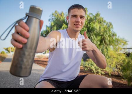 Un atleta sorridente tiene una bottiglia d'acqua e dà un pollice soddisfatto in su. Foto Stock