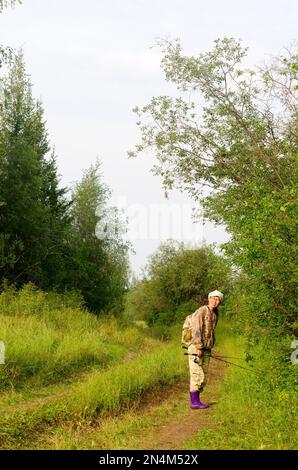 Pescatore asiatico Yakut in tuta protettiva e con uno zaino che tiene una canna da pesca gira sorridendo in piedi sul sentiero in erba. Foto Stock