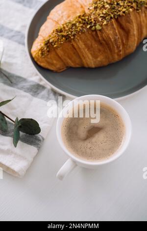 Primo piano di tazza di cappuccino con asciugamano da cucina, croissant e ramo di eucalipto sul tavolo Foto Stock
