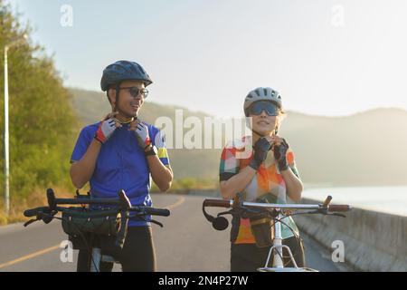 Coppia asiatica di ciclista con caschi e cinghie per il mento mentre si preparano per il loro giro in bicicletta intorno a un bellissimo lago mattutino. Foto Stock