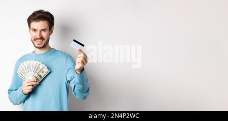 Bel giovane uomo sorridente e che offre il pagamento in contanti e senza contatto, mostrando denaro con carta di credito in plastica, in piedi su sfondo bianco Foto Stock