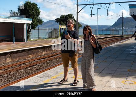 Stazione ferroviaria di St James Western Cape, Sud Africa. 2023. Turisti in possesso di telefoni cellulari, attendere un treno passeggeri sul binario di St James Stat Foto Stock