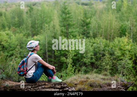 Yakut ragazza asiatica turista con zaino e cappello seduto sul bordo della montagna nella tundra selvaggia di Yakutia abbracciando le gambe riposando e guardando Foto Stock