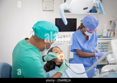 Un dentista africano controlla la salute orale del paziente mentre il suo assistente prepara gli strumenti di lavoro. Foto Stock