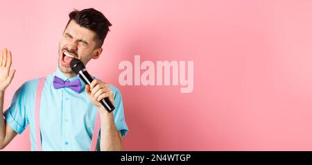 Concetto di festa ed eventi di festa. Il ragazzo divertente esegue una canzone, cantando il karaoke in microfono con la faccia spensierata, in piedi in bow-craving e sospenders sopra Foto Stock