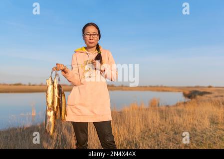 Yakut giovane ragazza gioiosa pescatore duro tenendo un sacco di pesce pescato luccio appeso al Pesce Stringer sullo sfondo del lago nel campo. Foto Stock