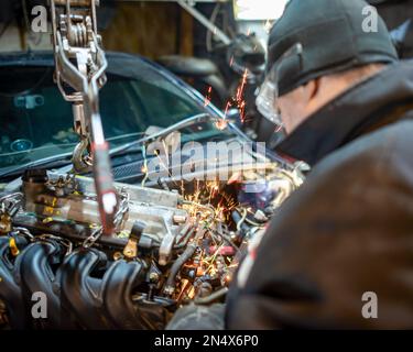 Un meccanico bespectacled dell'automobile nel garage funziona con le scintille che volano da sotto la sega che mette il motore nell'automobile. Foto Stock