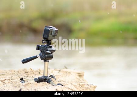 Action camera si trova su un piccolo treppiede, registrando video, sulla riva sabbiosa del fiume con erba e pioggia. Foto Stock