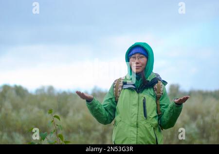 Yakut ragazza asiatica turista in una giacca cappello, cappuccio e occhiali, con uno zaino è sorridente, grandine, pioggia e neve tenendo le mani contro lo zaino Foto Stock