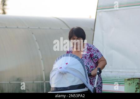 Donna anziana stanca Yakut porta un passeggino con un bambino sullo sfondo di serre con piante in giardino. Foto Stock