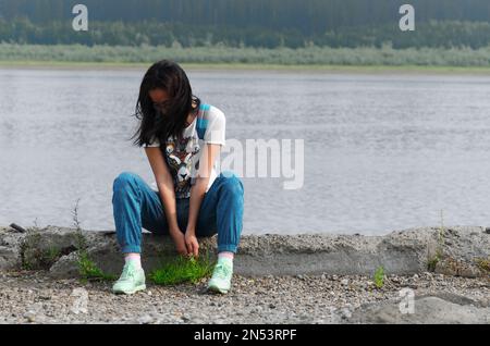 Una ragazza pensierosa si siede sul bordo di un vecchio molo sul fiume Viluy con le foreste di Yakutia nell'estremo nord della Russia che guardano il bo Foto Stock