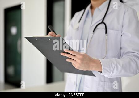 Immagine ritagliata di un medico professionista asiatico in camici bianchi nel corridoio dell'ospedale, scrivendo qualcosa sulla clipboard medica. Foto Stock