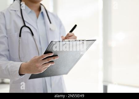 Immagine ritagliata di un medico femminile asiatico professionista in abito bianco in piedi nella sala esame, scrivendo qualcosa su una clipboard medica, o recor Foto Stock