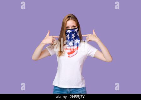 Giovane donna con maschera in forma bandiera america sul suo viso in t-shirt bianca mostra un gesto con le dita a se stessa, mentre in piedi su uno sfondo viola Foto Stock