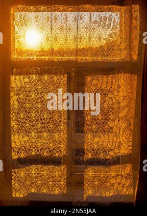 La luce del sole giallo brillante proveniente dal tramonto sull'orizzonte passa attraverso una finestra in una stanza rustica con una tenda a motivi bianchi e una cornice d'ombra. Foto Stock