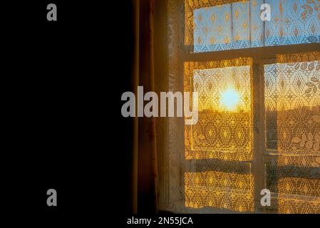 La luce del sole giallo brillante dal tramonto sull'orizzonte arriva attraverso la finestra nella stanza del villaggio con la tenda a motivi bianchi e la shad Foto Stock