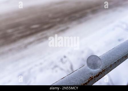 Una piccola palla di vetro si trova in inverno sulla ringhiera nella neve sullo sfondo di una strada fangosa. Foto Stock