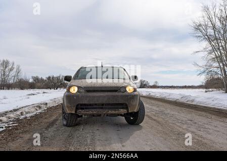 Un incrocio bianco sporco SUV si alza con i fari accesi prima di guidare su una strada invernale con la neve. Foto Stock