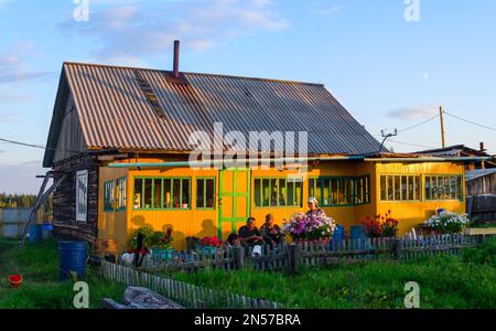 Il cortile di una casa privata Yakut con una recinzione vicino alla foresta e la gente seduta sulle scale tra i fiori al tramonto alla luce del sole in sha Foto Stock