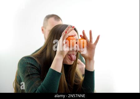 Ragazza giovane mettere tangerini agli occhi e tirato fuori lingua bloccato e lei torce e rende i volti in background un uomo su uno sfondo bianco verde maglione lungo capelli scherzo folgorante intorno a grimacing Foto Stock