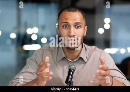 Primo piano ritratto uomo con un microfono professionale, afro-americano registrazione audio e video podcast seduto in studio, chiamata di un serio uomo d'affari parlando e chiacchierando con i colleghi. Foto Stock