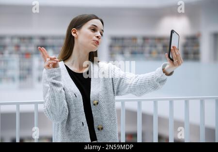 Selfie, biblioteca e donna con segno di pace per i social media, influencer blog post o di studio aggiornamento per lo stile di vita gen z. Emoji mani persona o studente Foto Stock