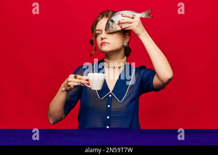 Giovane donna stravagante con tazza di caffè e pesce crudo su sfondo rosso brillante. Gusto delizioso e strano. Vintage, stile retrò. Colori complementari Foto Stock