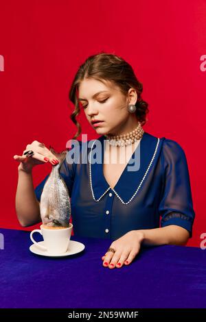 Tempo del tè. Giovane donna stravagante con tazza di caffè e pesce crudo su sfondo rosso brillante. Gusto delizioso e strano. Vintage, stile retrò. Cibo pop art Foto Stock