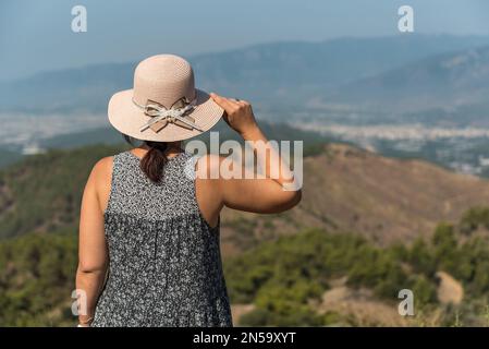 Giovane donna che guarda in lontananza, belle montagne sullo sfondo, pensando profondamente, indossando abito e cappello causale, natura, vita lenta, viaggio Foto Stock