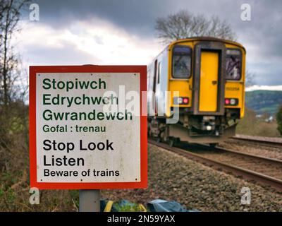 Un treno supera un segnale di avvertimento che dice "Stop Look Listen" in gallese e inglese ad un passaggio pedonale di una linea ferroviaria. In una giornata trascorsa in inverno Foto Stock