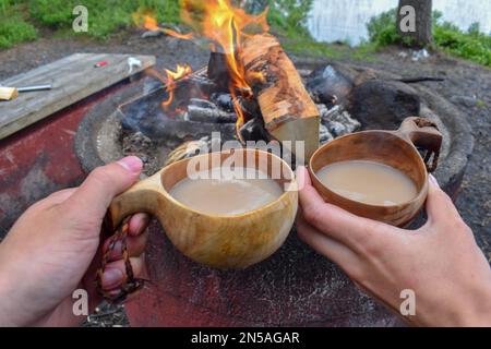 Tazza gialla con bevanda calda su ceppo di legno vicino falò all'aperto, spazio per testo. Stagione Campeggio. Nel nord della Svezia. Foto Stock