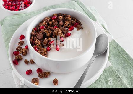 Ciotola di yogurt greco con granola di avena al cioccolato e semi di melograno su tavola di marmo bianco. Colazione sana Foto Stock
