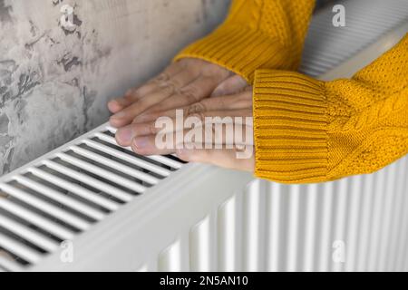 Primo piano dell'uomo che scalda le mani in maglione giallo sul riscaldatore a casa durante le fredde giornate invernali. Maschio scaldarsi le braccia sopra il radiatore. Concetto Foto Stock