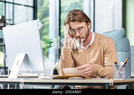 Uomo sconvolto in ufficio leggere il messaggio sulla carta, uomo d'affari ha ricevuto la busta di posta con notifica e cattive notizie, seduto sul posto di lavoro con il computer. Foto Stock