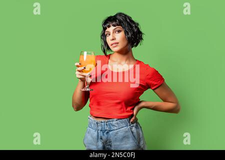 Ritratto di una giovane bruna, ventenne, che tiene un bicchiere di succo d'arancia appena spremuto. Indossa jeans e una t-shirt rossa ed è po Foto Stock
