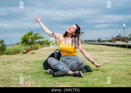 bella giovane donna latina seduta a prendere il sole con gli occhi chiusi e il contenuto seduta a gambe incrociate sull'erba sorridente, vestita di giallo, braccia fuori Foto Stock