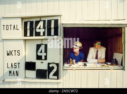 Gli scorer che osservano la partita di cricket locale del villaggio dal tabellone segnapunti tradizionale Foto Stock