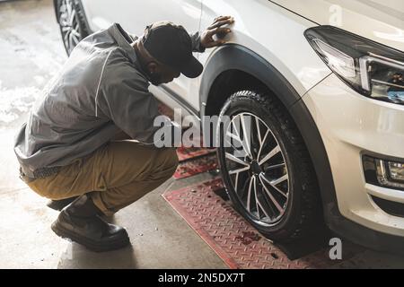 Meccanico afro-americano che controlla la geometria di una ruota in un negozio di riparazione di automobile, vista laterale del colpo pieno. Foto di alta qualità Foto Stock