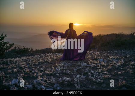 Tramonto viola abito donna montagne. Ascesa del mistico. tramonto sulle nuvole con una ragazza in un lungo vestito viola. Nel prato c'è un prato Foto Stock