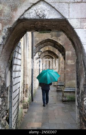 Uomo che cammina sotto la pioggia con un ombrello sotto gli archi dei contrafforti volanti sul lato della Cattedrale di Winchester, Winchester, Hampshire, Inghilterra, Regno Unito Foto Stock