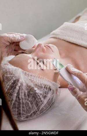 Cosmetologist, estetician che fa il trattamento facciale con la spatola ultrasonica alla donna giovane, trattamento di scrubber della pelle del viso con la spatola ultrasonica, procedura di pulizia facciale nel salone di bellezza. Foto Stock