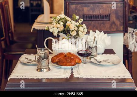 Un set tavolo per bere tè in stile retrò dei tempi dell'URSS - torte al forno, bicchieri sfaccettati in metallo stand, teiera, vaso con fiori, tovaglioli con pizzo. Foto Stock