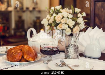 Un set tavolo per bere tè in stile retrò dei tempi dell'URSS - torte al forno, bicchieri sfaccettati in metallo stand, teiera, vaso con fiori, tovaglioli con pizzo. Foto Stock