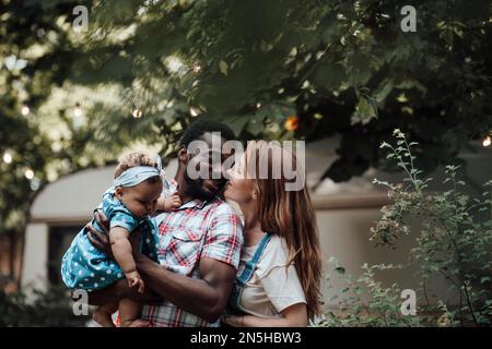 La famiglia di corse miste con una bambina dalla pelle scura passa il tempo insieme a abbracci e baci nel parco camper. L'uomo afroamericano la sua moglie e li della pelle fiera Foto Stock