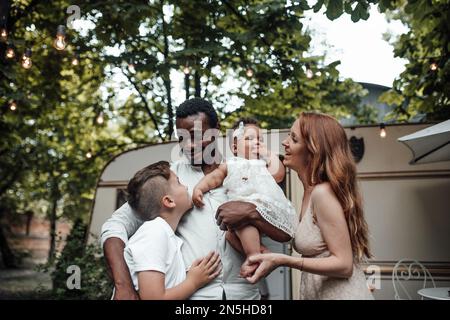La famiglia di corse miste con il figlio bianco e la bambina swarthy bambino trascorrono del tempo insieme nel parco camper all'aperto. Uomo afroamericano con la sua bella pelle wi Foto Stock