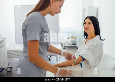 Giovane donna brunetta in un cappotto bianco rende le procedure mediche. Un'infermiera nei guanti e negli abiti medici prende il sangue o mette un contagocce sul paziente. MED Foto Stock