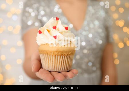 Donna che tiene gustoso cupcake per San Valentino contro le luci sfocate, primo piano Foto Stock